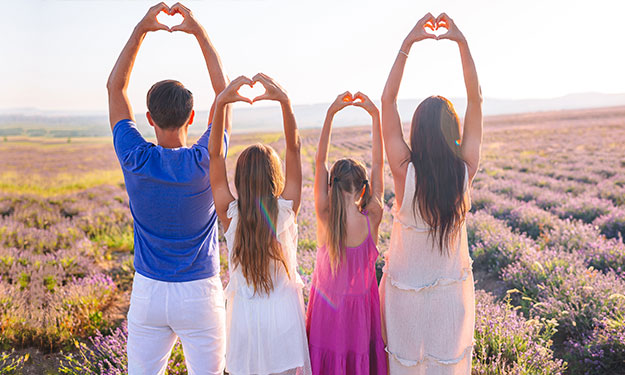A family of 4 people, standing in the middle of lavender field. They stand back side to the viewer. Their hands are reaching up, and showing the heart symbol with their fingers.