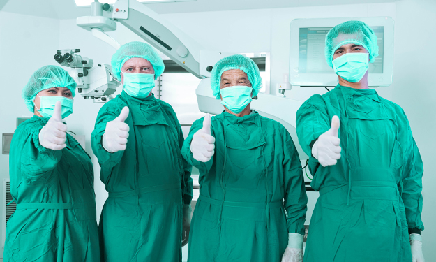 Four nurses stand in an operating theatre, dressed in sterile garments, showing their thumbs in a positive sign.