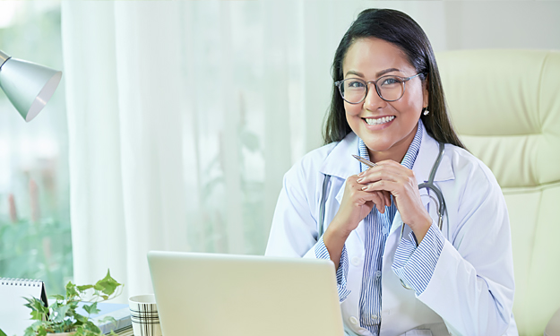 A smiling doctor with glasses and a white coat sits at her desk in front of a laptop and looks at the viewer with her beautiful blue eyes.